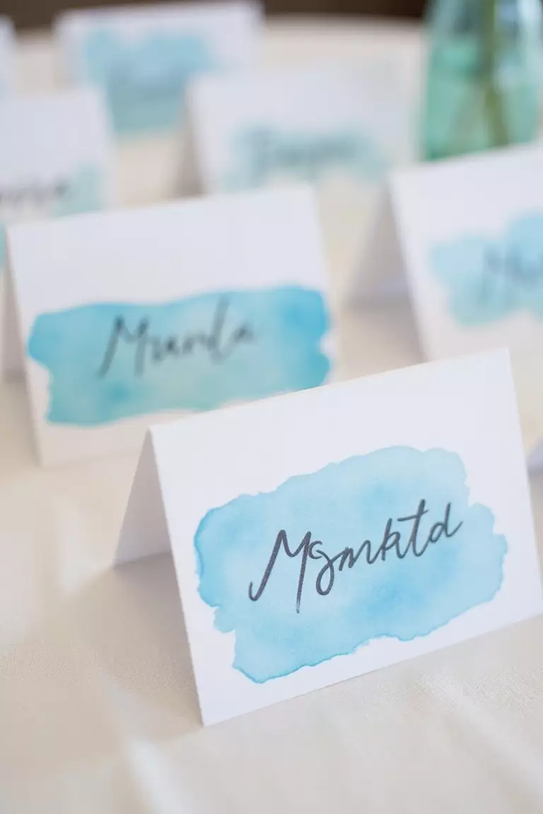 Blue Watercolor Place Cards A professional photo, similar to a photo in a wedding magazine, of hand-painted blue watercolor place cards with calligraphy displayed on a white linen tablecloth.