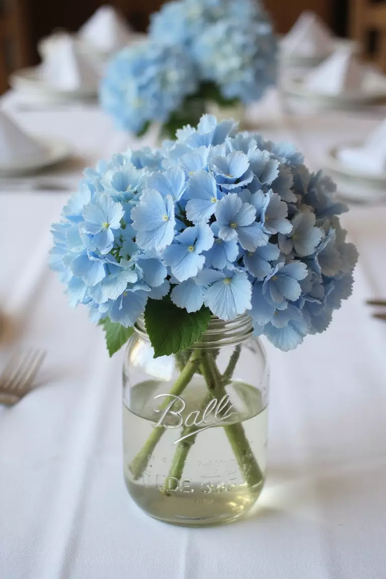 Mason Jar Centerpieces With Blue Hydrangeas A professional photo, similar to a photo in a wedding magazine, of rustic mason jars filled with lush blue hydrangeas as elegant centerpieces on a white linen tablecloth.