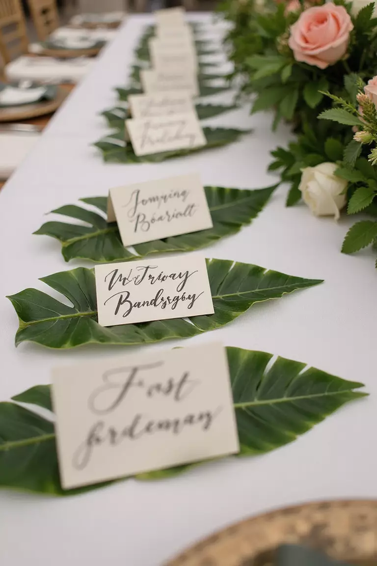 Palm Leaf Place Cards A professional photo, similar to a photo in a wedding magazine, of handwritten place cards elegantly displayed on small, glossy palm leaves at a tropical wedding reception table setting.