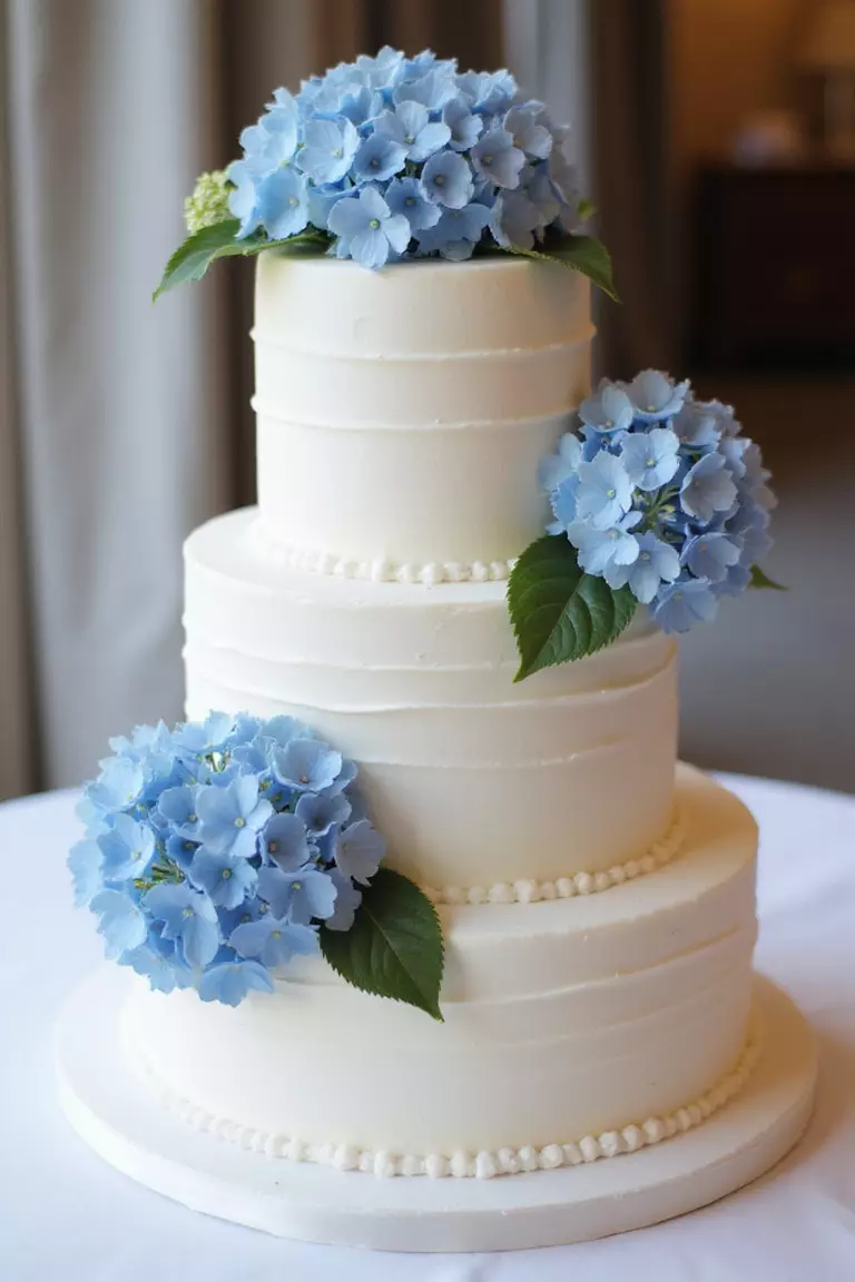 Cake Decorated With Fresh Blue Hydrangeas A professional photo, similar to a photo in a wedding magazine, of a three-tiered white wedding cake elegantly decorated with fresh blue hydrangea blooms between layers.