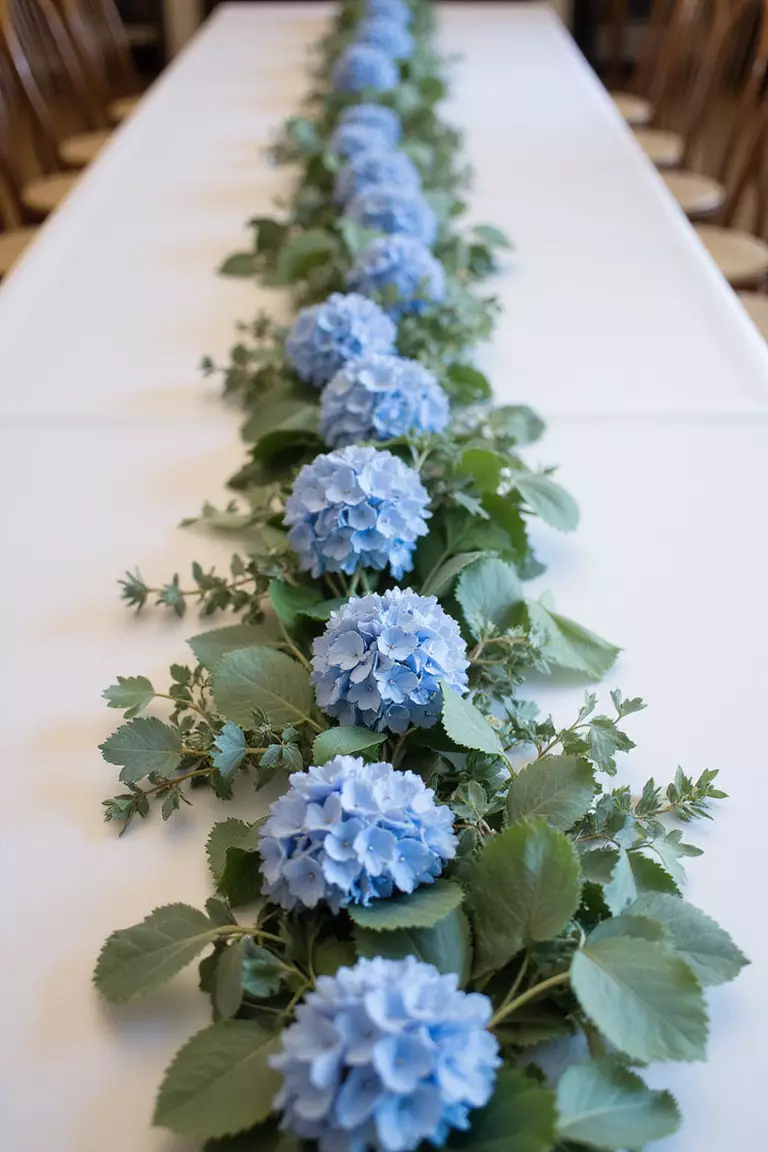 Hydrangea Garland For Head Table A professional photo, similar to a photo in a wedding magazine, of a lush garland of blue hydrangeas and eucalyptus running along the length of a white head table.