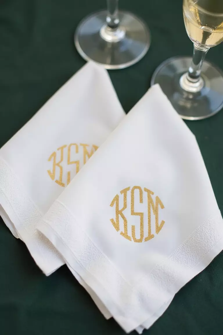 Gold Foil Monogrammed Cocktail Napkins A professional photo, similar to a photo in a wedding magazine, of cocktail napkins with elegant gold foil monograms, folded perfectly beside champagne glasses on a dark green surface.