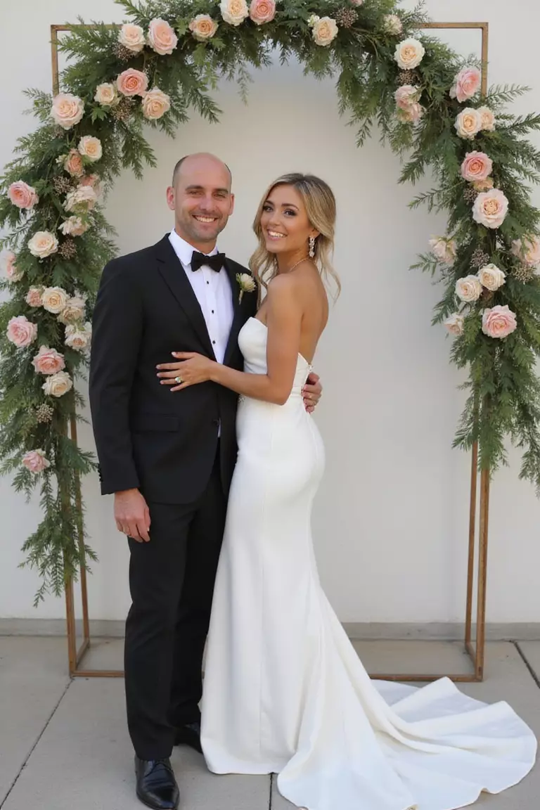 Unique Wedding Attire A professional photo, similar to a photo in a wedding magazine, of a bride smiling radiantly in an elegant white jumpsuit, standing beside her partner under a floral arch.