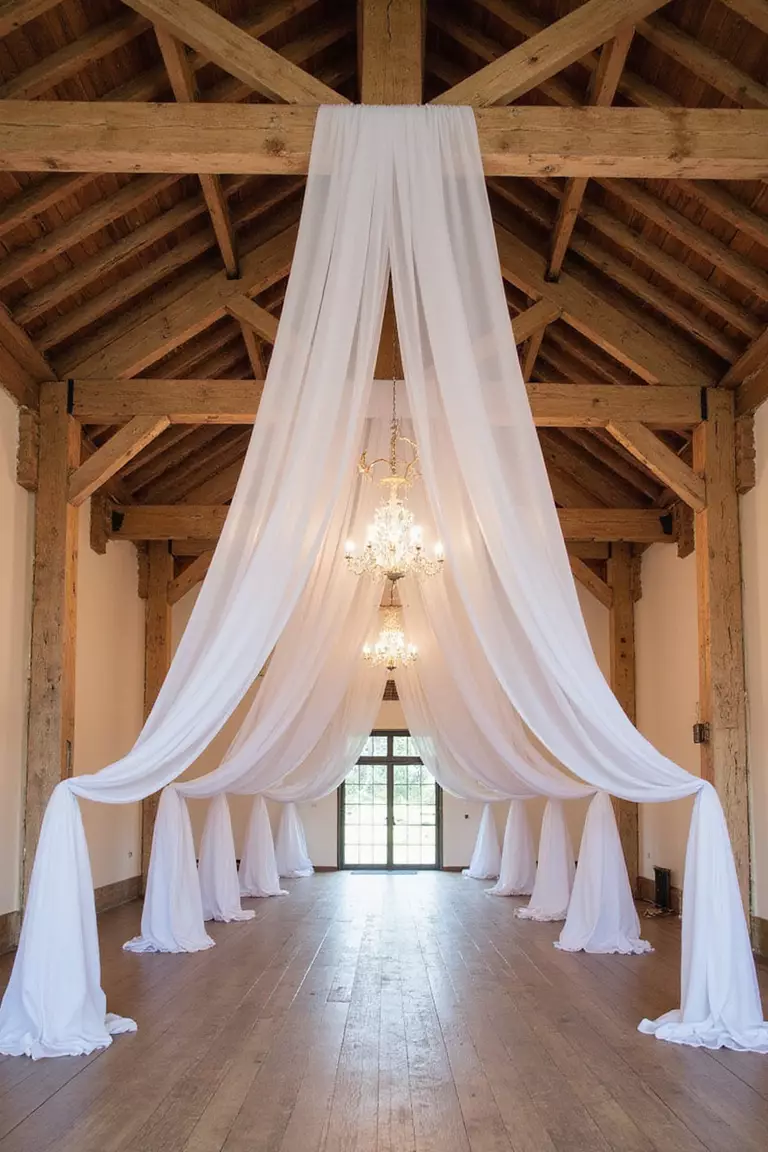 Lightweight Fabric Draping A professional photo, similar to a photo in a wedding magazine, of sheer white fabric draped elegantly from the beams of a rustic barn ceiling for a wedding reception.