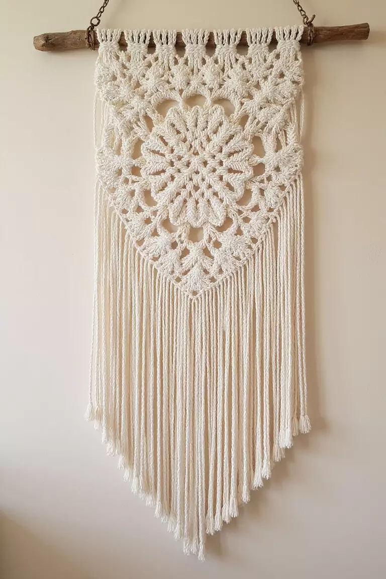 Macrame Wall Hanging A professional photo, similar to a photo in a wedding magazine, of a large, intricate ivory macrame wall hanging with fringe details, mounted on a rustic wooden dowel against a soft beige wall.