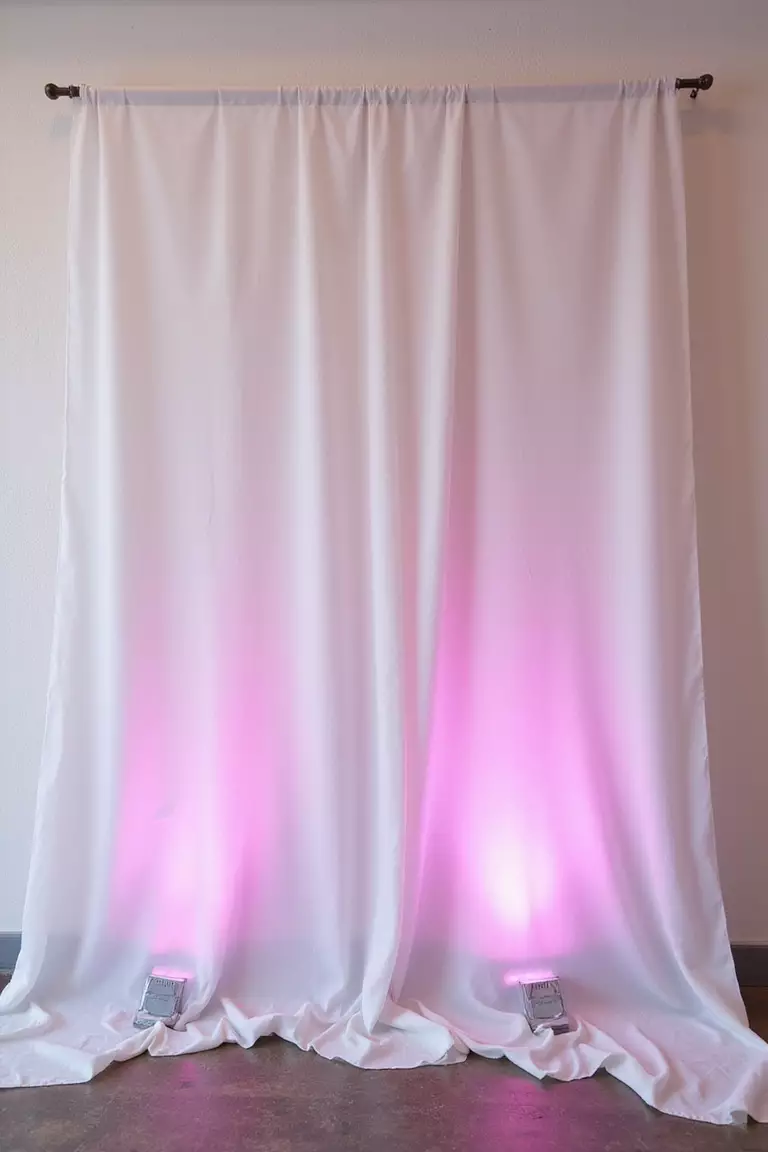 Fabric Draping With Uplighting A professional photo, similar to a photo in a wedding magazine, of soft white sheer fabric elegantly draped from ceiling to floor against a wall, with soft pink uplighting from the base.