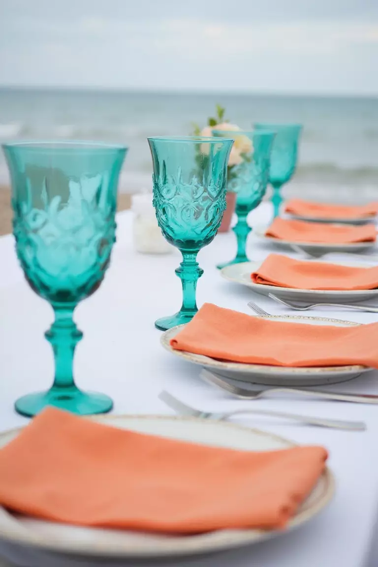 Turquoise And Tangerine Zest A professional photo, similar to a photo in a wedding magazine, of turquoise glassware and tangerine-colored napkins on a white linen tablecloth at a beach wedding reception. Ocean background.