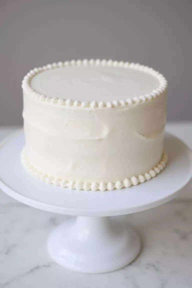 Classic White Elegance A professional photo, similar to a photo in a wedding magazine, of a round, single-tier white buttercream cake with simple piped pearl borders, on a clean white cake stand.