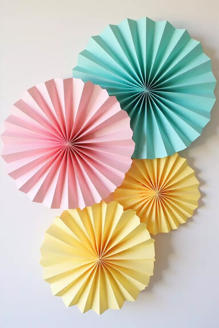 Paper Fan Rosettes A professional photo, similar to a photo in a wedding magazine, of colorful paper fan rosettes in pink, teal, and yellow, arranged in an overlapping cluster on a white wall.