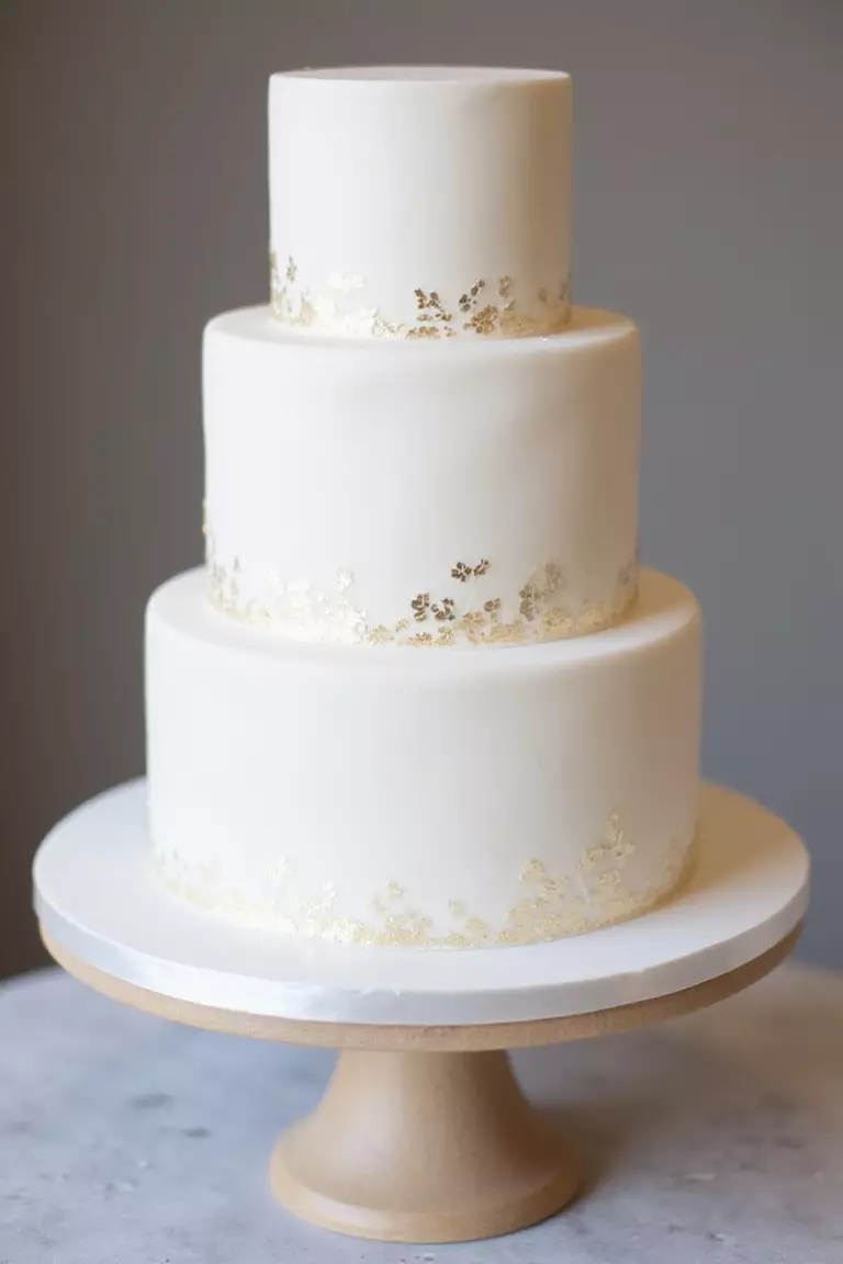 Metallic Accents Cake A professional photo, similar to a photo in a wedding magazine, of a sleek white fondant cake with delicate metallic gold leaf accents applied sparingly around the base.