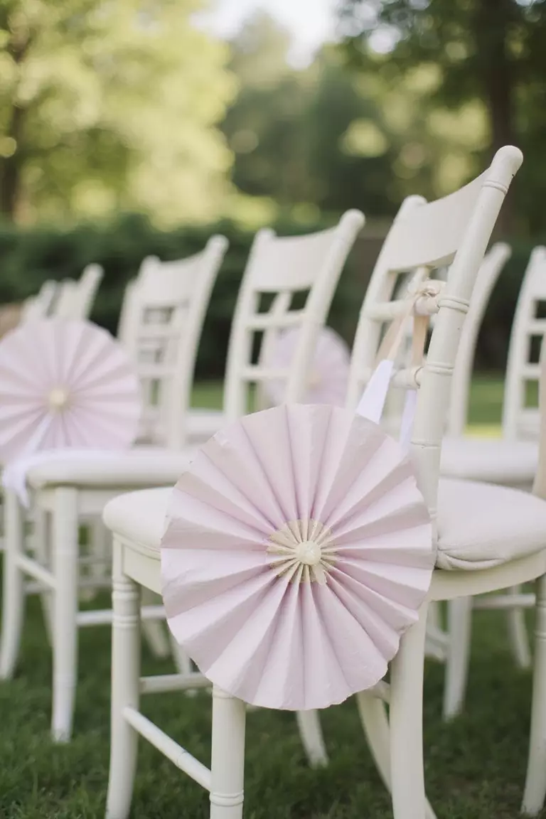 Give Fans As Wedding Favors A professional photo, similar to a photo in a wedding magazine, of pretty paper hand fans placed on each chair at an outdoor summer wedding ceremony setup.