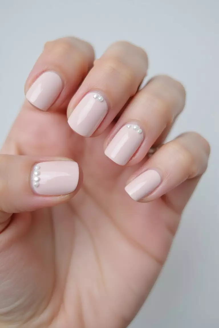 Short and Sweet: Chic Designs for Natural Nails Close-up of a woman's hand with short, natural-looking nails featuring a neutral pink polish with tiny pearl accents against a light, clean background.