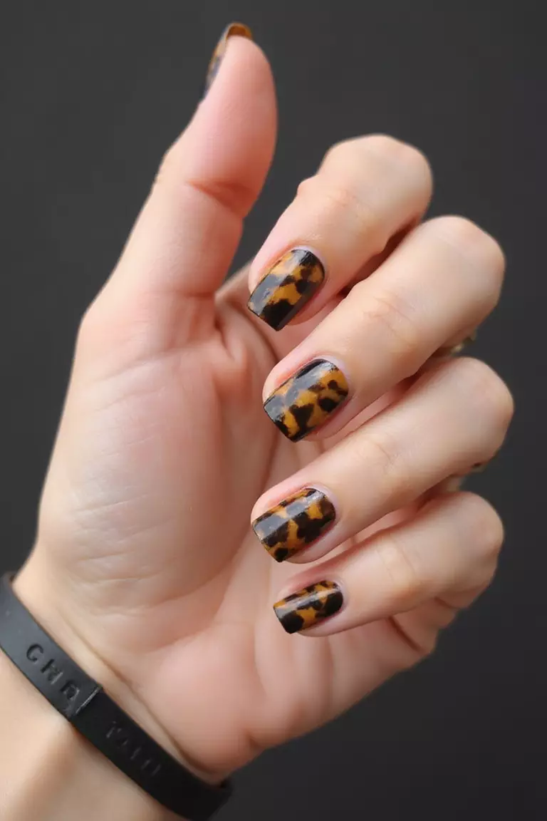 Tortoise Shell Close-up of a woman's hand with five fingers featuring short nails with tortoiseshell pattern in amber, brown, and black, creating a stylish neutral design.