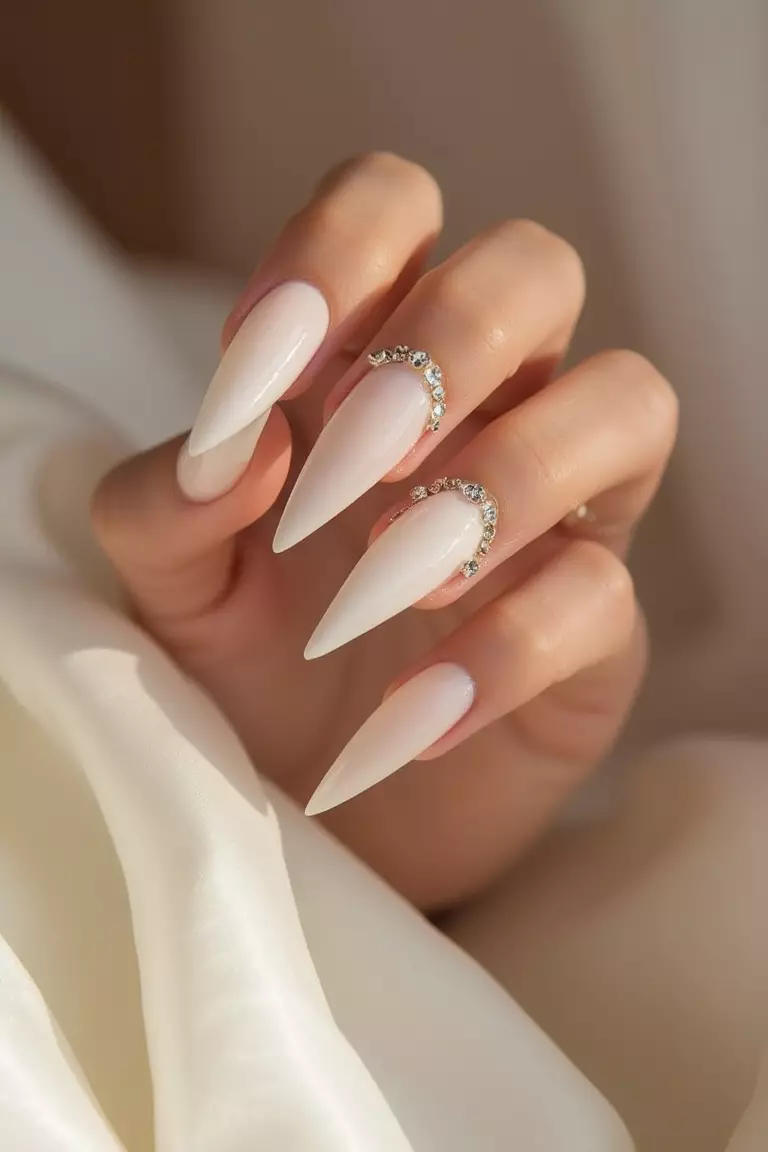 Close-up of a woman's hand showing sharp stiletto nails in pearl white with delicate crystal accents at the base, captured with soft lighting against satin fabric.