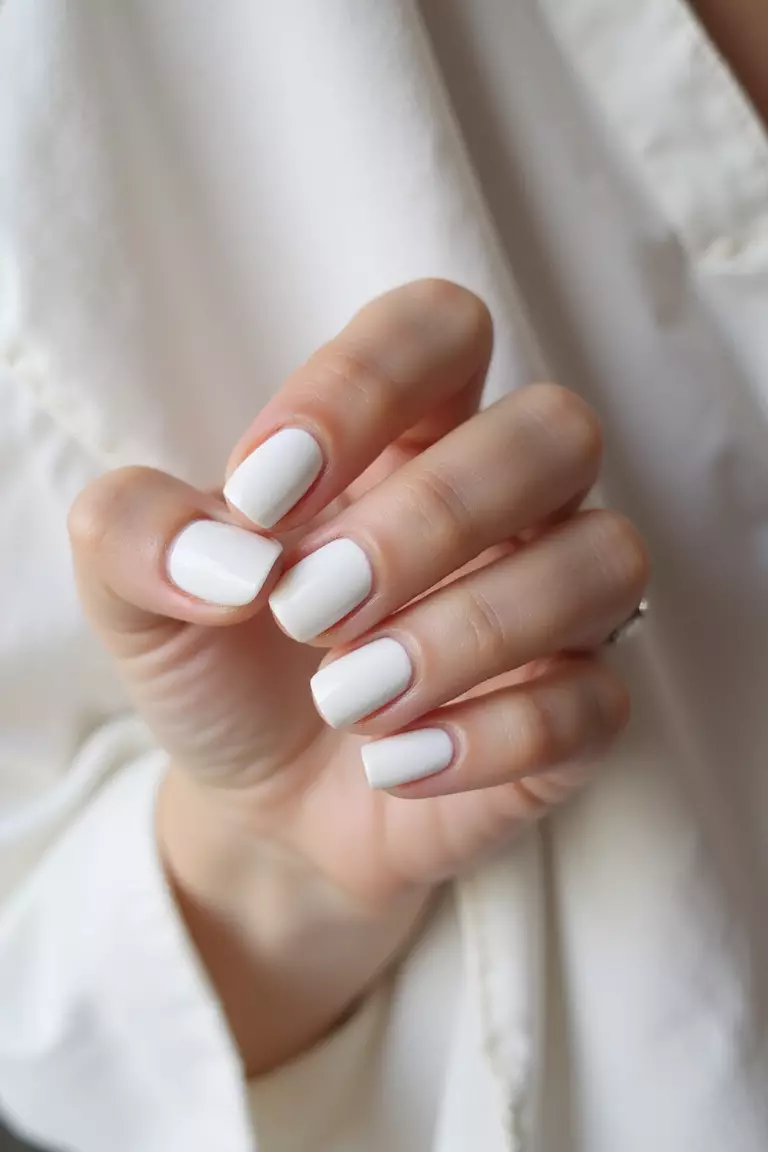 Close-up of a woman's hand displaying milky white nails with subtle matte finish, photographed against soft fabric, natural light emphasizing the clean, minimalist design.