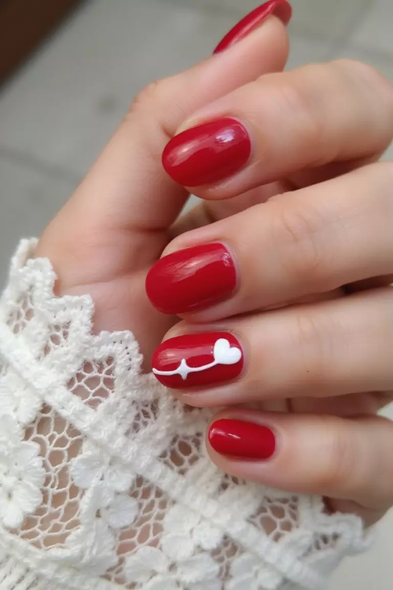 Close-up of a woman's hand with red nails, one featuring a small white heart accent design, posed gently against white lace.