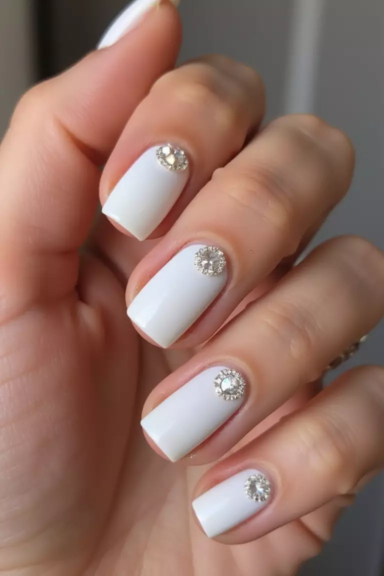 Diamond Accent Nails Close-up of a woman's hand with five fingers featuring white nails with tiny diamond-like crystals placed strategically, catching light with every movement.