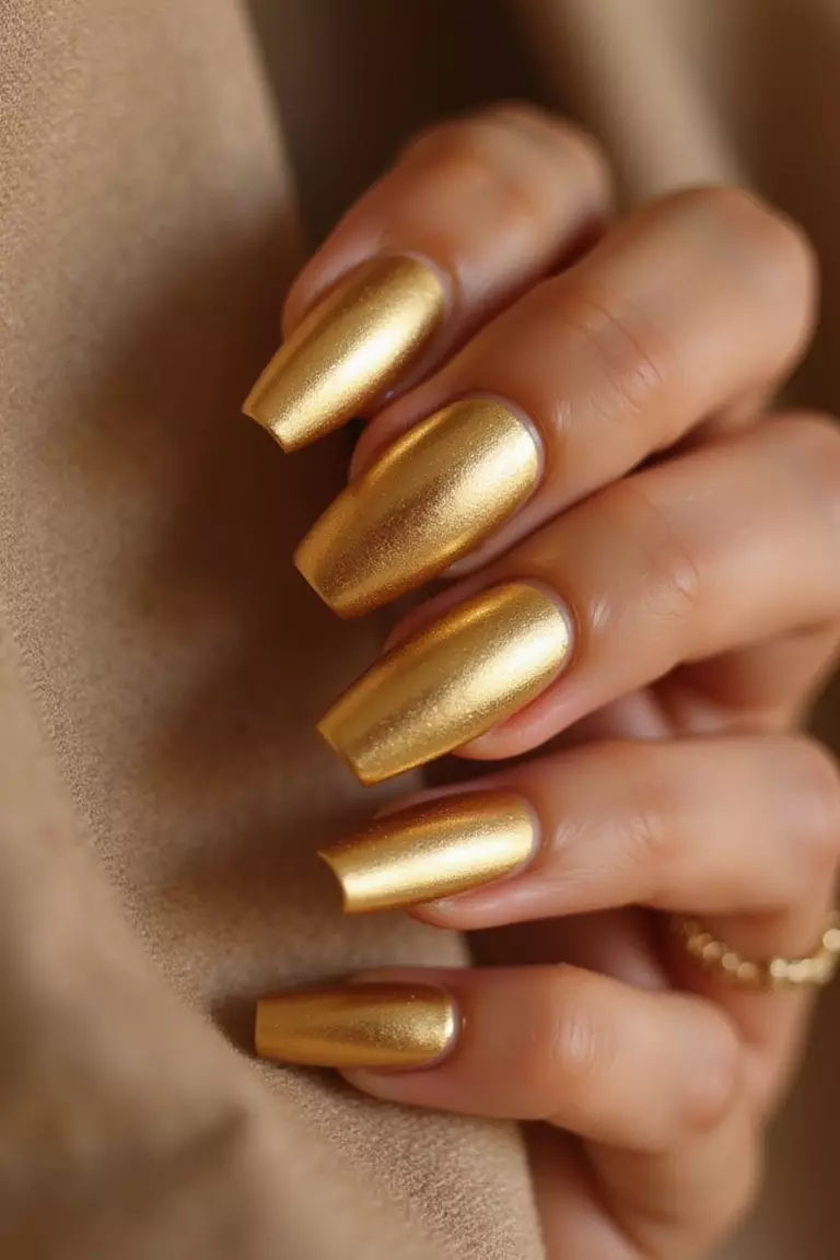 Close-up of a woman's hand with full gold metallic nails reflecting warm light, photographed against neutral fabric to highlight the rich, luxurious shine.