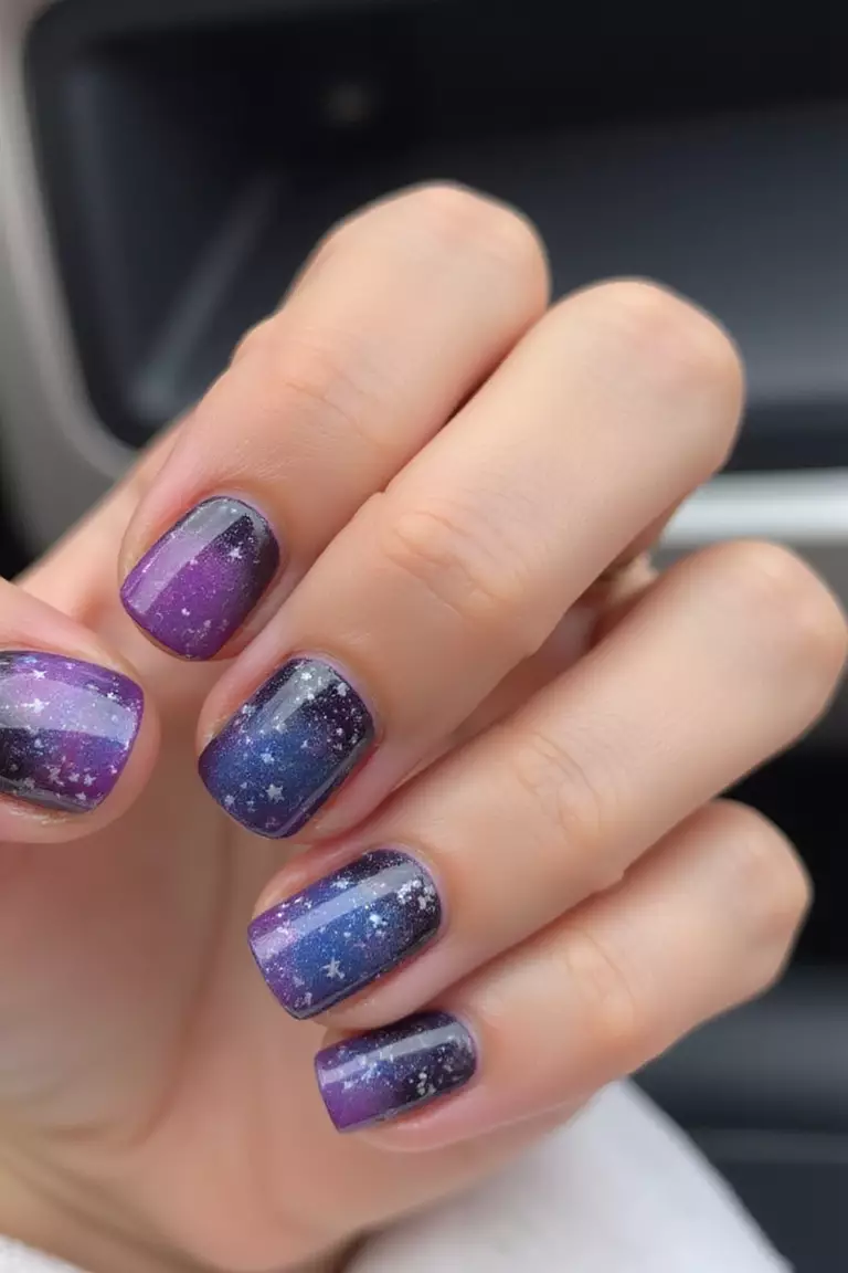 Out of this World Close-up of a woman's hand with five fingers featuring short nails with galaxy-inspired design showing purples, blues, and specks of white resembling stars.