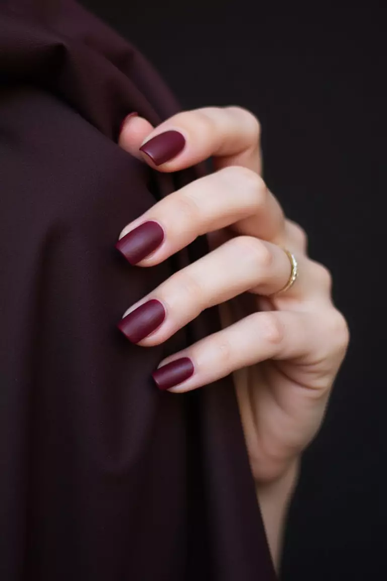 Close-up of a woman's hand with deep wine-red matte nails that look velvety and sophisticated, posed elegantly against dark fabric.