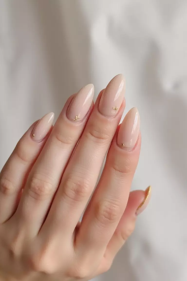 Almond Allure: Flattering Shapes for Elegant Hands Close-up of a woman's hand with almond-shaped nails in a soft champagne color with delicate gold cuticle accents against a light, airy background.
