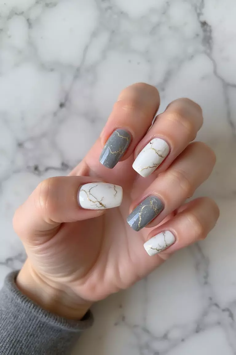 Marble Magic: Sophisticated Stone-Inspired Nail Art Close-up of a woman's hand with white and gray marble-effect nails featuring delicate gold veining and a glossy top coat against a marble surface background.