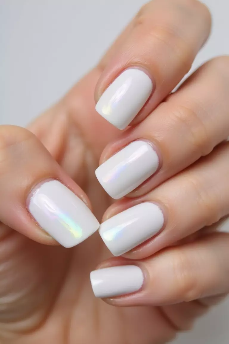 Close-up of a woman's hand with pearlescent white nails featuring a high-gloss, slightly iridescent finish that resembles glazed icing, captured in bright light against neutral background.
