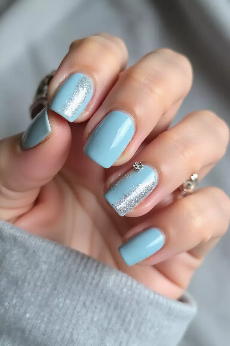 Something Blue: A Touch of Tradition with Modern Flair Close-up of a woman's hand with pale blue nails featuring subtle silver glitter accents and a tiny rhinestone on the ring finger against a soft fabric background.
