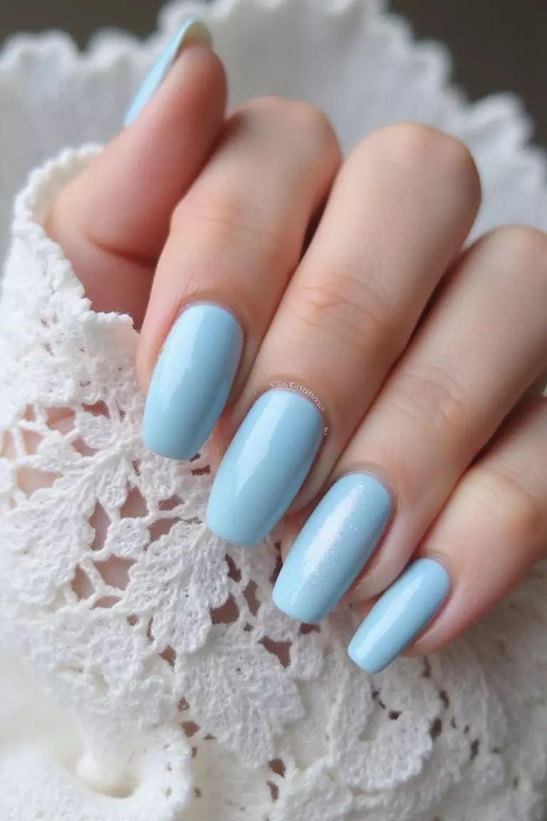 Close-up of a woman's hand with pale blue nails featuring a subtle pearlescent finish, photographed against white lace to highlight the delicate cool tone.