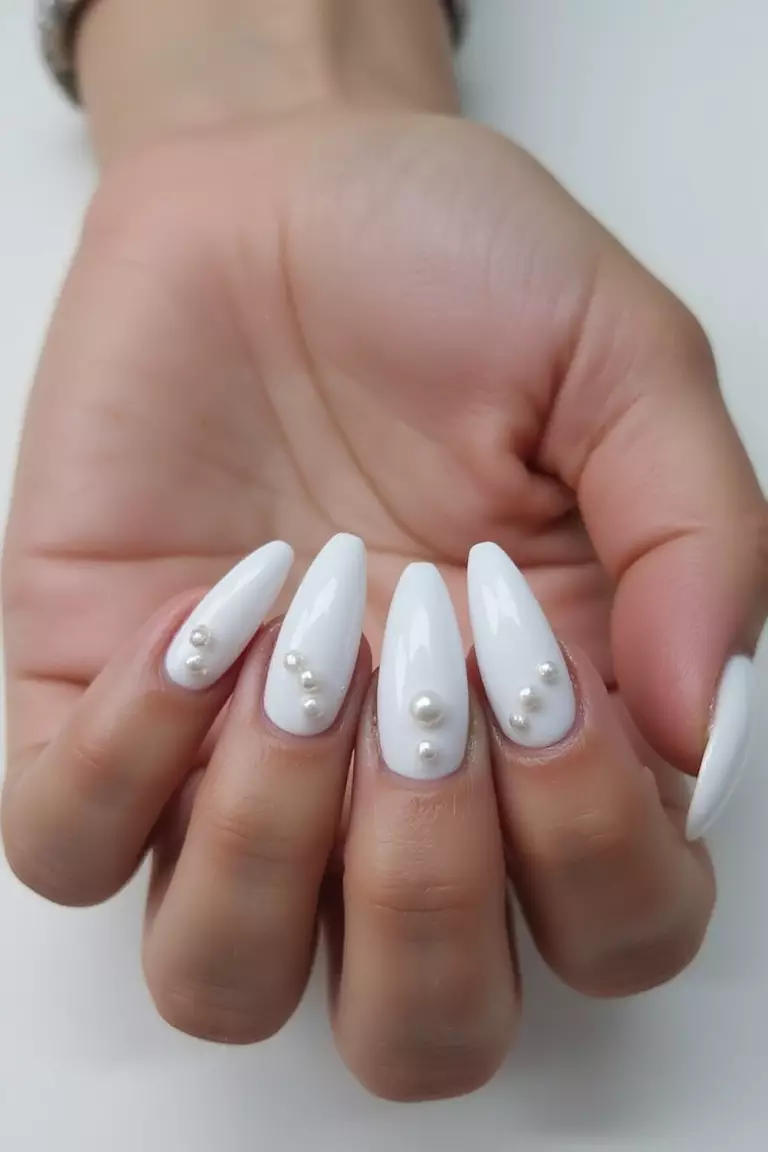 Long Acrylic Pearl Nails Close-up of a woman's hand with five fingers featuring white long, almond-shaped acrylic nails with embedded pearl elements throughout the design.