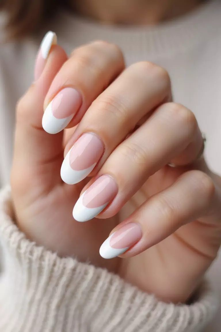 Close-up of a woman's hand with almond-shaped nails featuring classic white tips on a pale pink base, natural lighting highlighting the glossy finish against soft fabric.