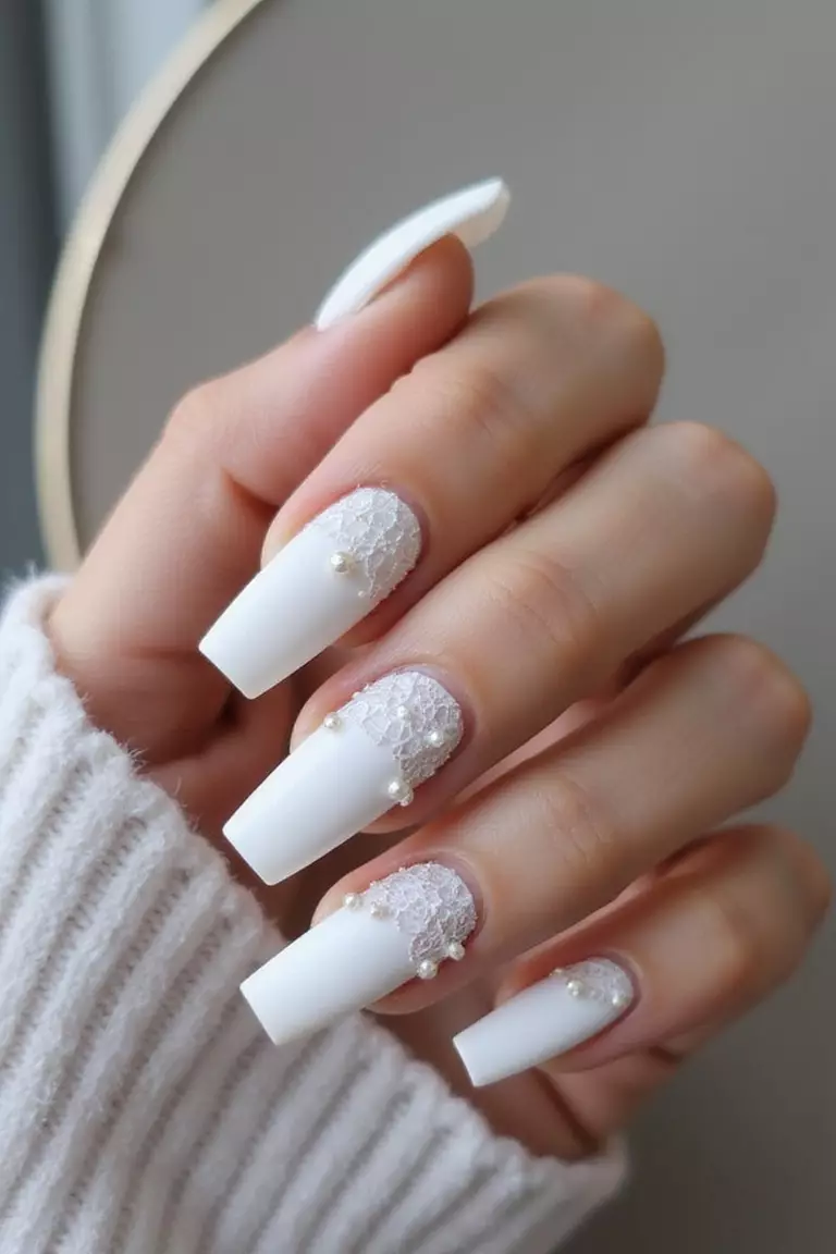 Coffin Nail Couture: Bold Statements for Fashion-Forward Brides Close-up of a woman's hand with long coffin-shaped nails in matte white with intricate lace-like details and tiny pearls against a modern, stylish background.