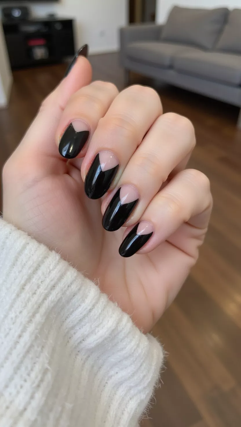 close-up shot of a woman’s hand with five fingers showing nails with a sharp, angular V-shaped chevron pattern painted in glossy black over a clear or nude base, living room background.
