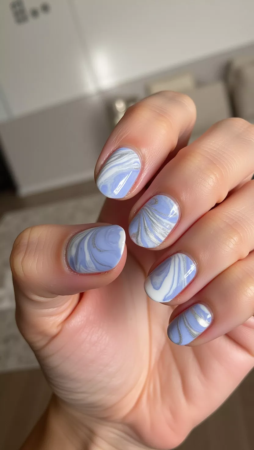 close-up photo of a woman’s hand with five fingers showing nails with a soft periwinkle blue base marbled with swirls of white and thin silver striping, room background