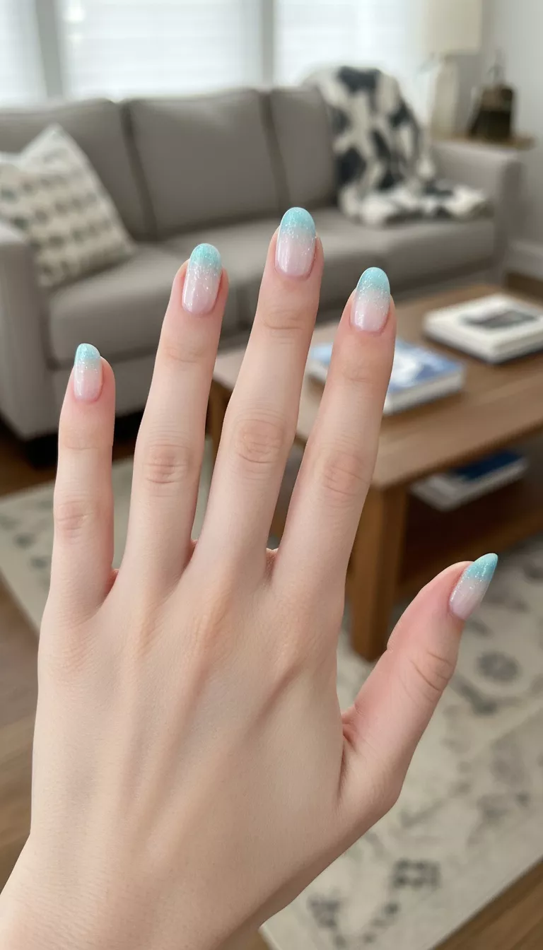 close-up shot of a woman’s hand with five fingers showing nails with a gradient effect transitioning from a sheer white at the cuticle to a crystalline icy blue shimmer at the tip, living room background.