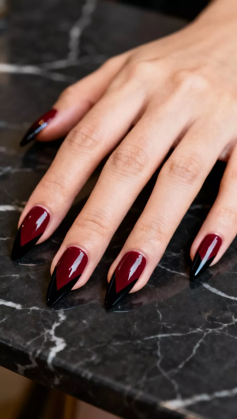 close-up shot of a woman’s hand with five fingers showing nails with a glossy burgundy base and a sharp black V-shape applied at the base of the nail, a dark marble table background.