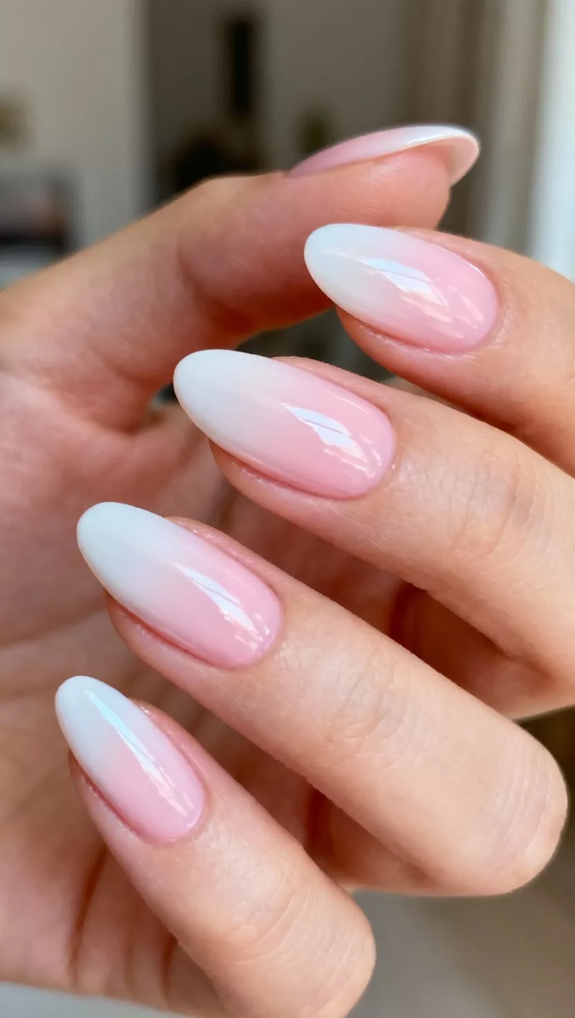 Icy Pink Stiletto Nails close-up shot of a woman’s hand with five fingers showing almond shaped nails with an extreme glossy light icy pink polish that appears nearly white in certain lights, room background.