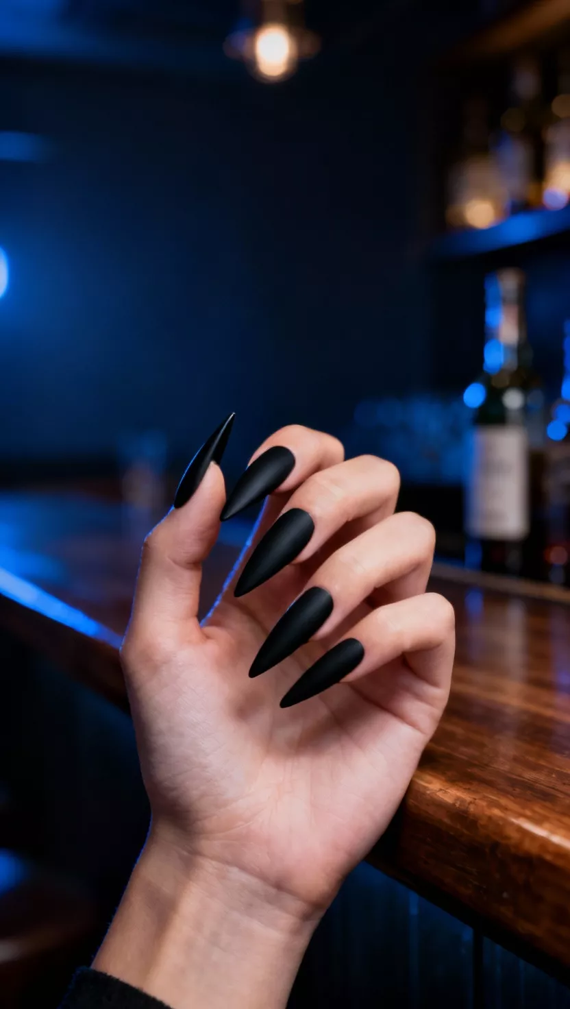 Black Stiletto Nails with Matte Finish close-up shot of a woman’s hand with five fingers showing nails with long, sharp stiletto shaped nails painted in a striking matte black, moody bar setting background.