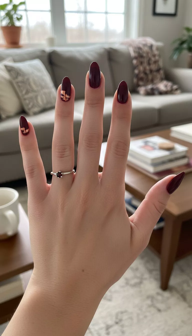 close-up shot of a woman’s hand with five fingers showing nails coated in a rich, deeply pigmented burgundy cream polish, with small, abstract swipes of metallic copper polish near the cuticles, living room background.