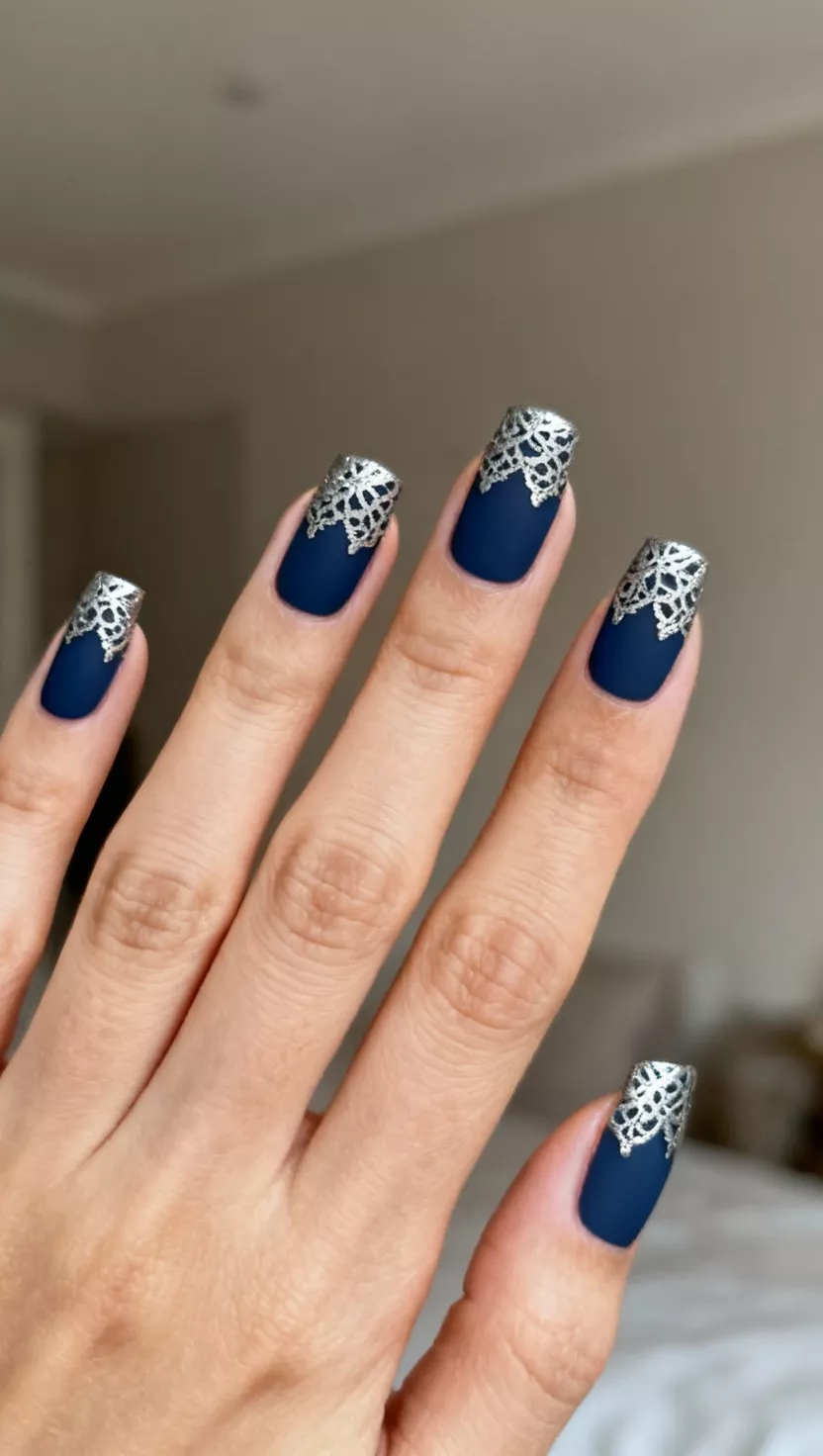 Midnight Blue with Silver Stamping close-up shot of a woman’s hand with five fingers showing nails with a matte midnight blue base color, featuring intricate, almost lace-like silver stamping patterns across the tip of each nail, room background.