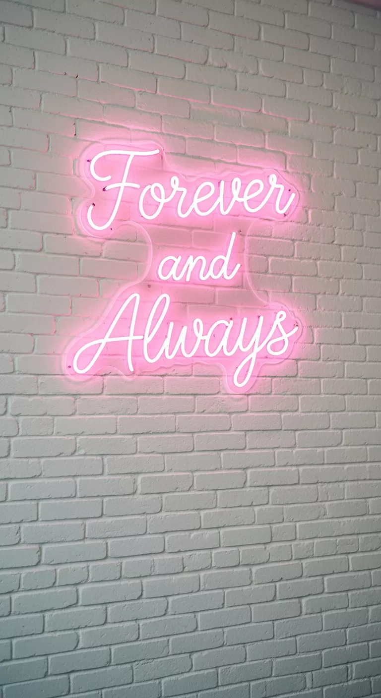 A professional photo, similar to a photo in a wedding magazine, of a white brick wall decorated with a pink neon sign displaying 'Forever and Always' in cursive script, acting as a modern, playful backdrop for guest photos at a wedding reception.