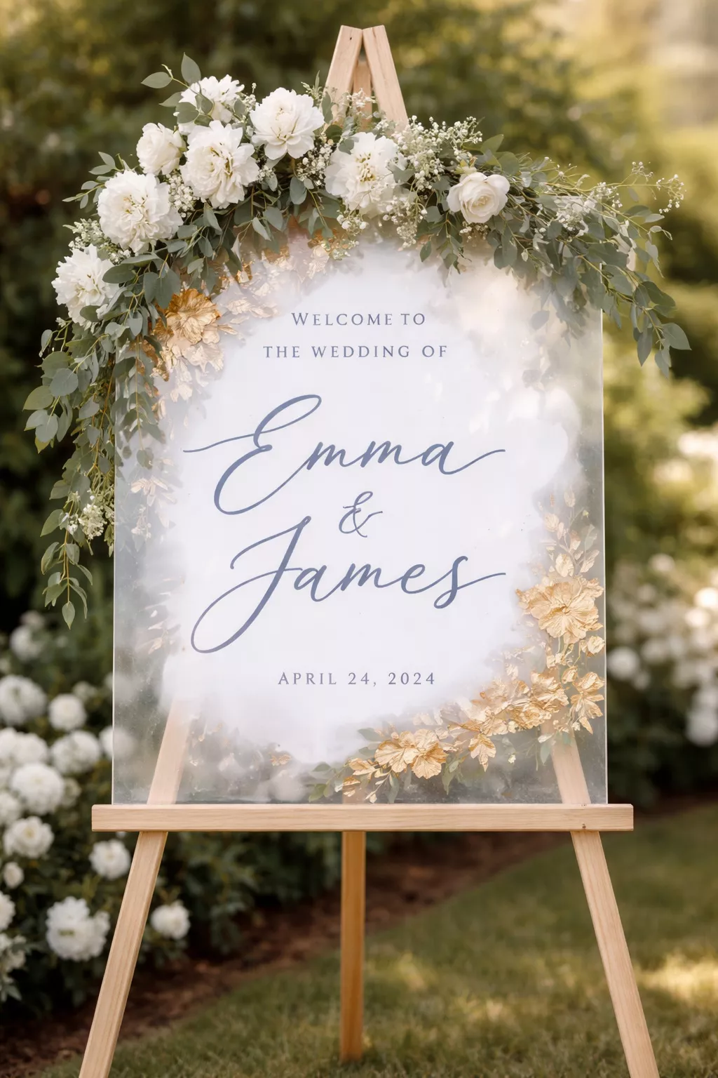 A realistic photo of an acrylic wedding welcome sign with dusty blue calligraphy and gold leaf accents on a wooden easel garlanded with white flowers