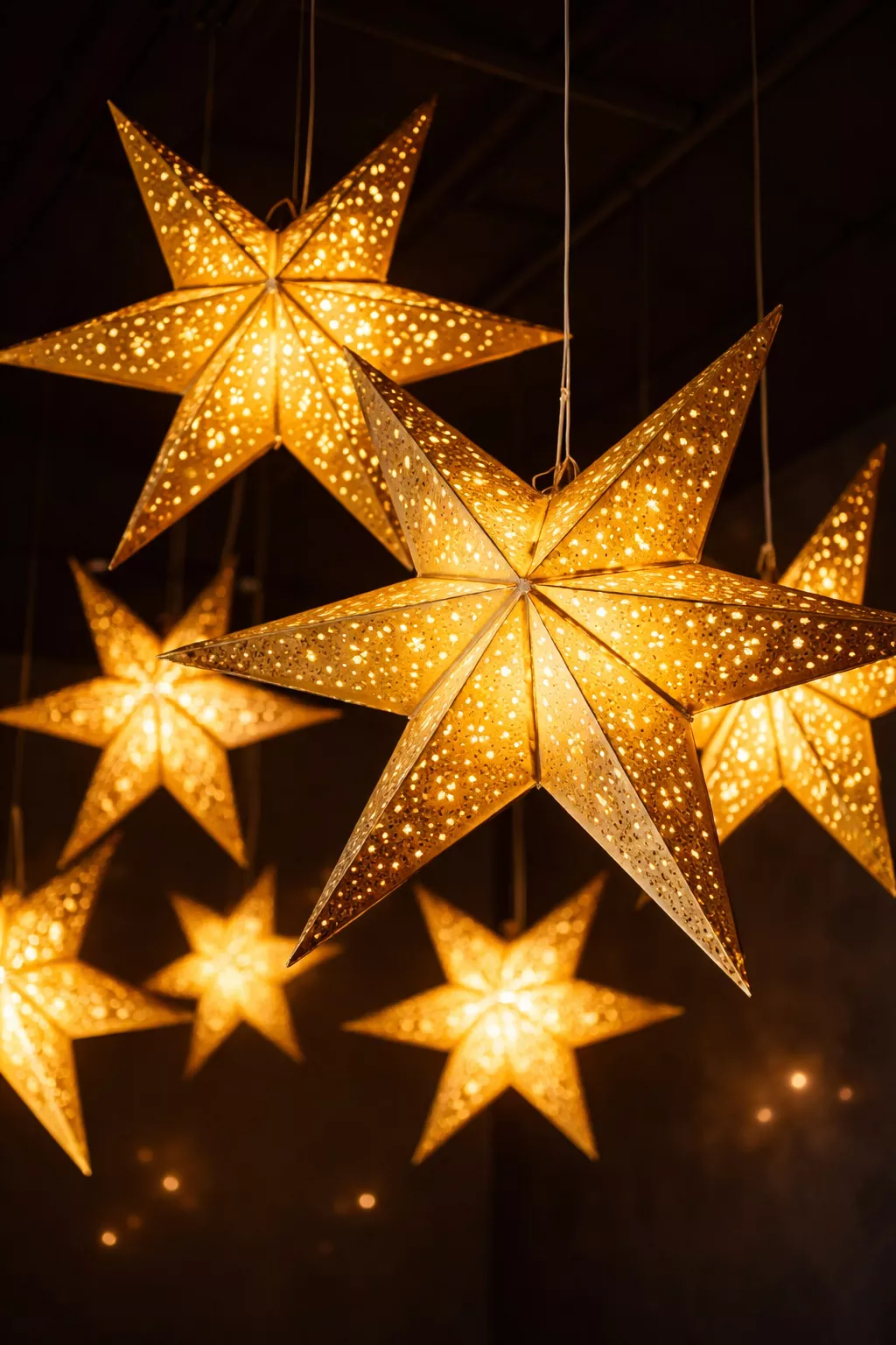 A realistic photo of gold foil star-shaped paper lanterns hanging from a dark ceiling, glowing with internal warm white lights.