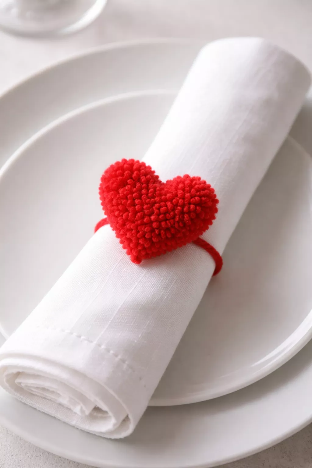A realistic photo of a white cloth napkin with a small red yarn pom-pom shaped like a heart serving as a napkin ring, resting on a white plate.