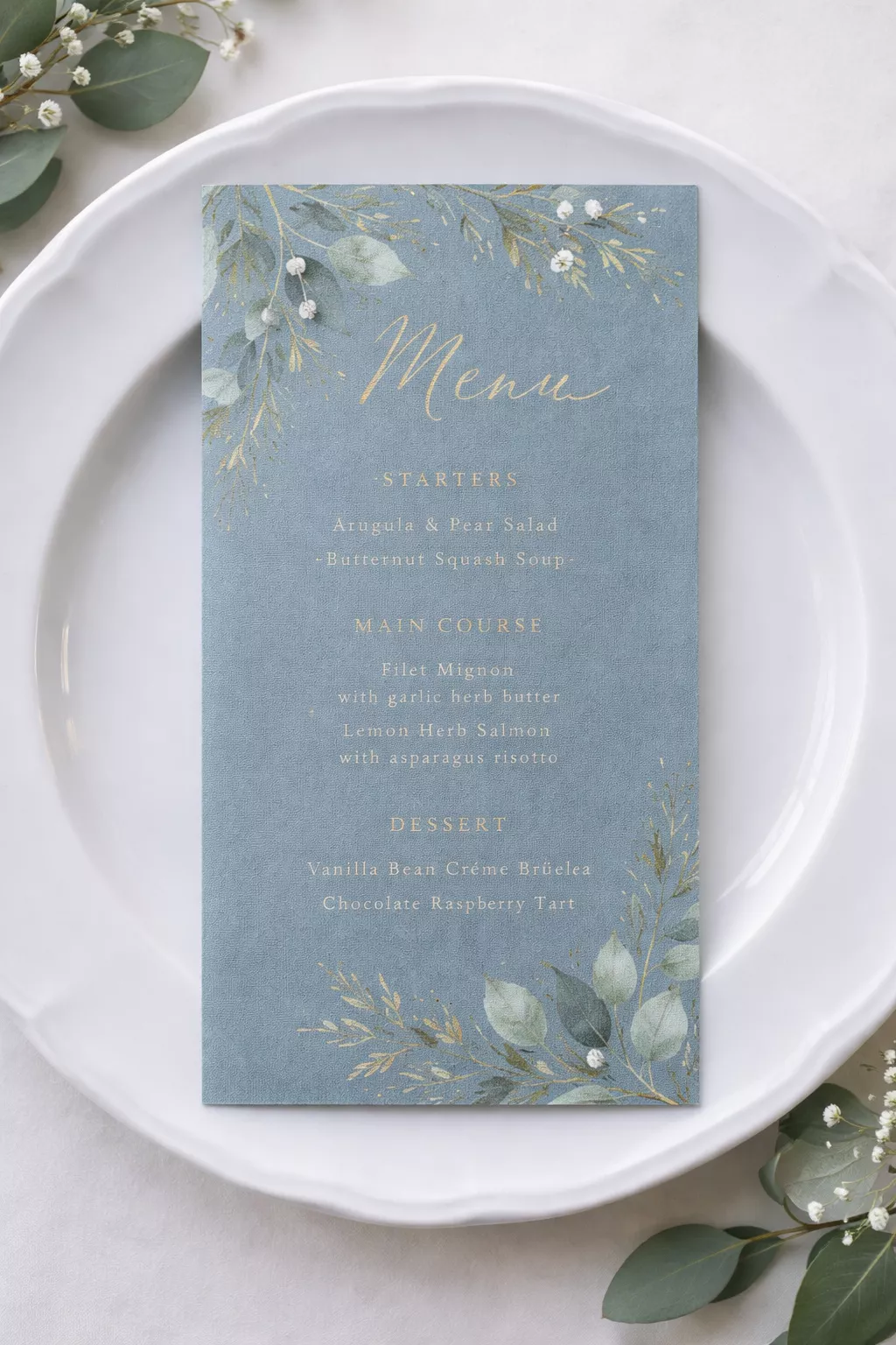 A realistic photo of a textured dusty blue paper menu card with gold ink and greenery illustrations placed on a white porcelain plate