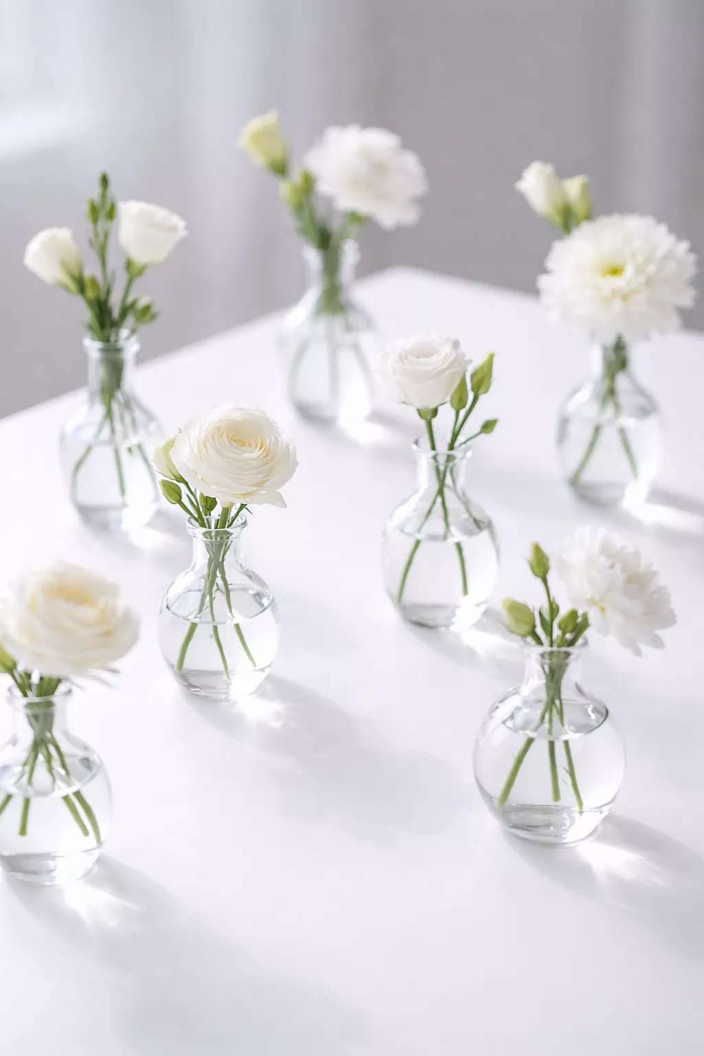 A realistic photo of several small clear bud vases each holding a single white flower on a clean white table.