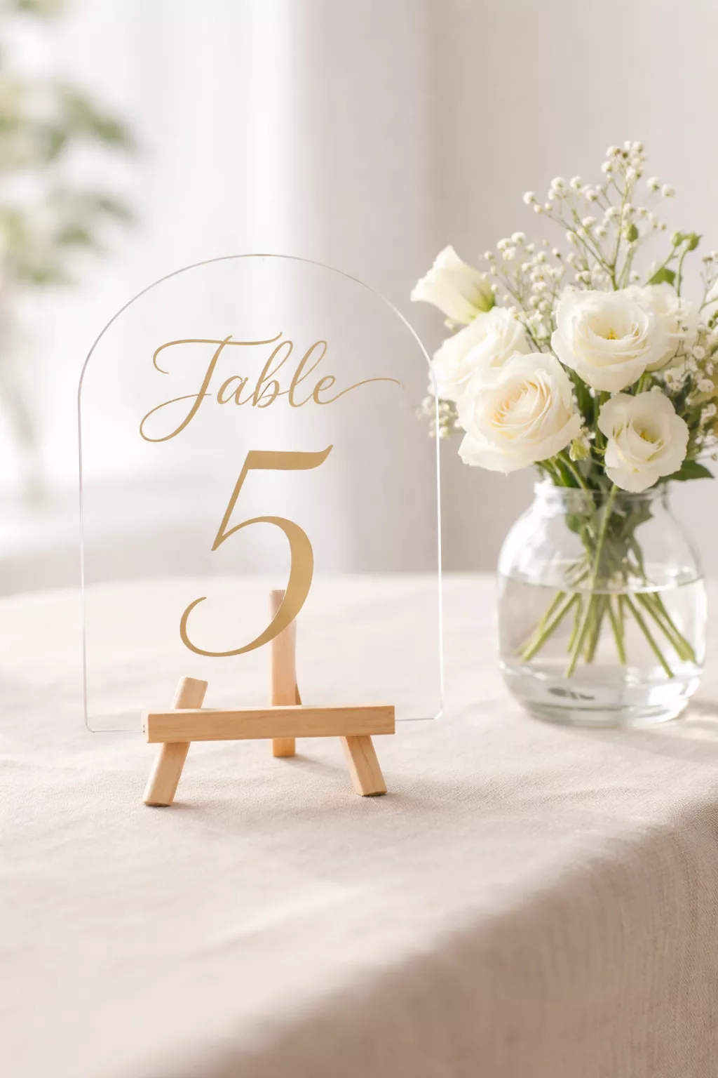 Personalized Table Numbers A realistic photo of a clear acrylic table number arch with elegant gold calligraphy lettering, sitting on a miniature wooden easel next to a small glass vase of white flowers.