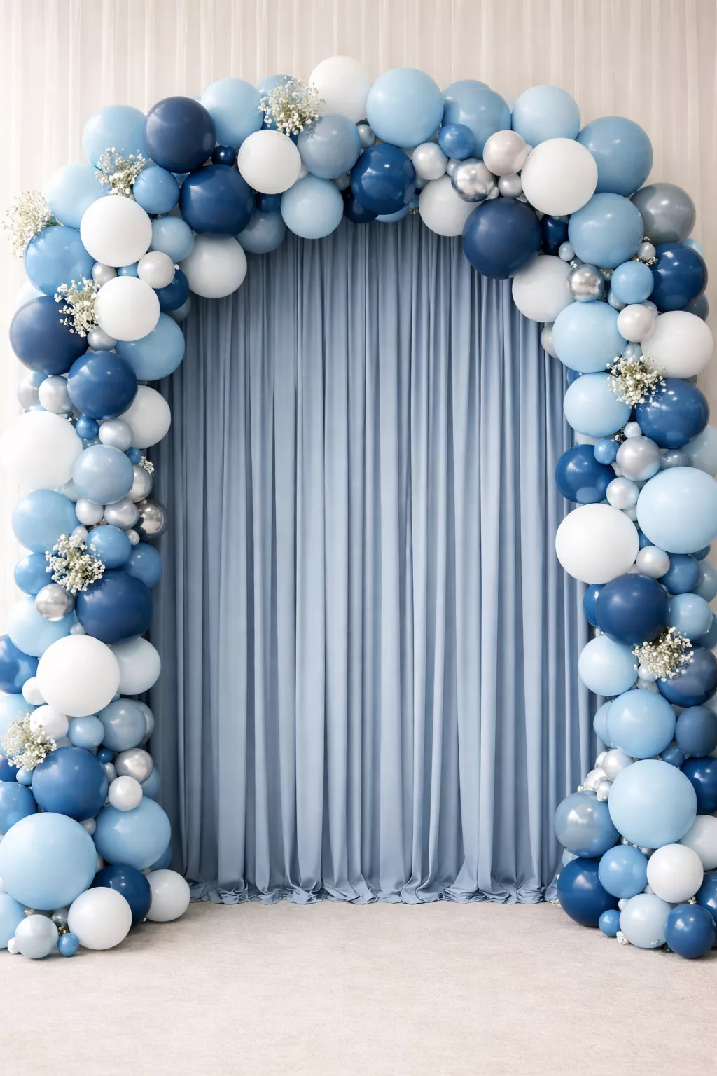 A realistic photo of a photo booth backdrop featuring vertical dusty blue fabric streamers and a large balloon arch in shades of blue and white