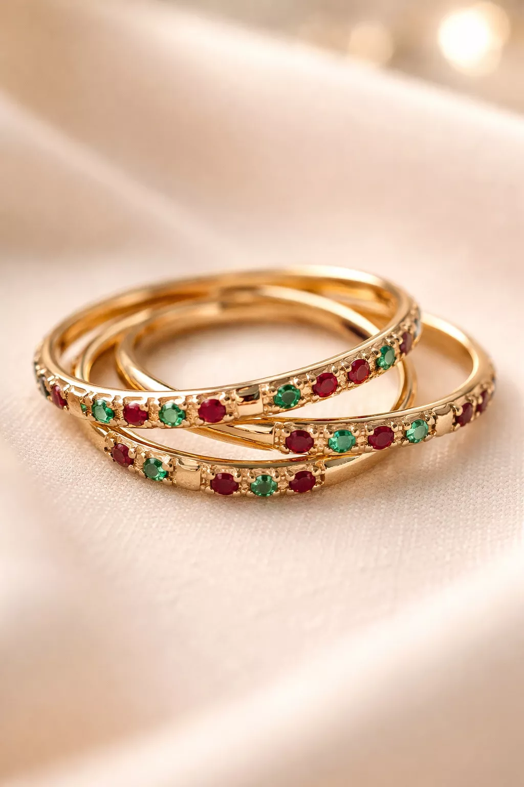 A realistic photo of three thin gold interlocking wedding bands set with small green emeralds and red rubies.