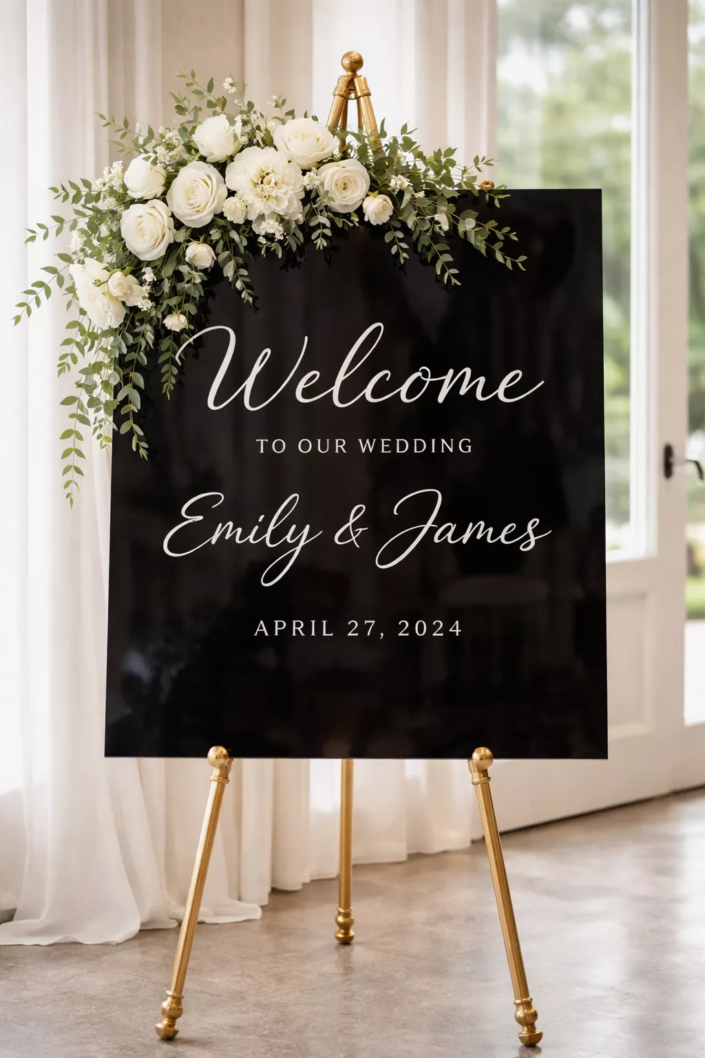 A realistic photo of a large black acrylic welcome sign with white script lettering, placed on a gold easel and decorated with a cluster of white flowers.